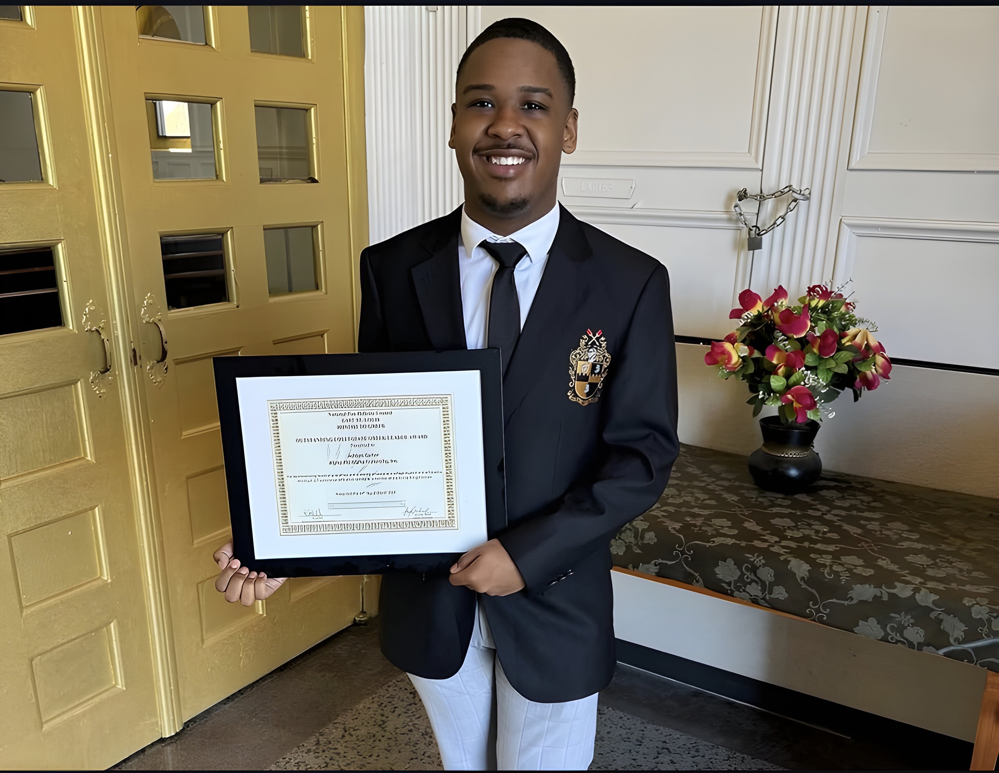 Ashton holding a framed award certificate in a black blazer with fraternity crest