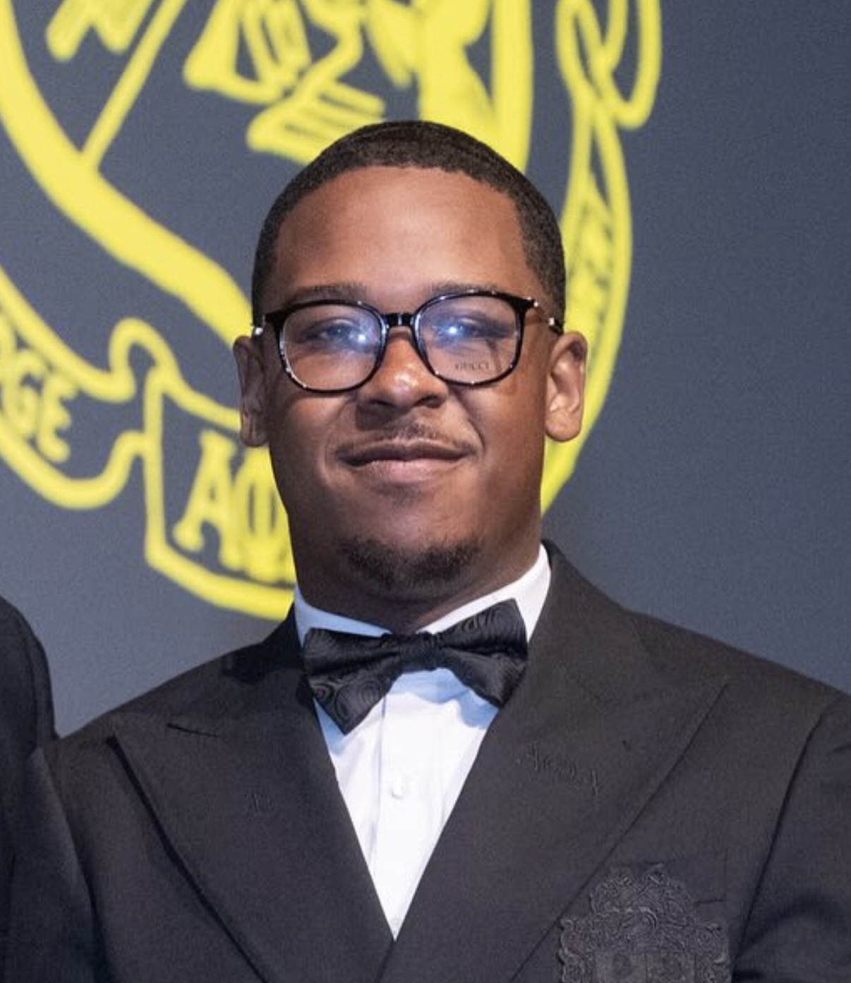 Ashton in a tuxedo with bow tie, smiling in front of a fraternity crest